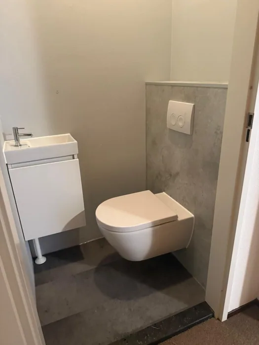 Hangtoilet tegen een voorzetwand in betonlook met witte drukplaat en een fonteinmeubel ernaast
