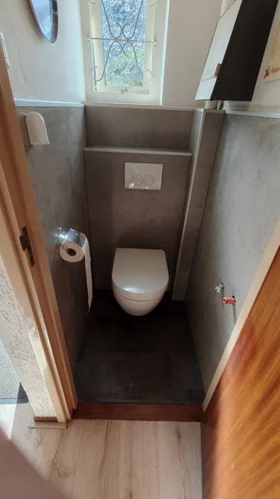 Toiletruimte na de renovatie: hangtoilet, voorzetwand met drukplaat en grijze betonlook panelen onder het raampje