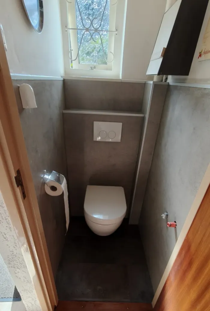 Na: dezelfde toiletruimte met hangtoilet, voorzetwand met drukplaat en grijze betonlook wandpanelen