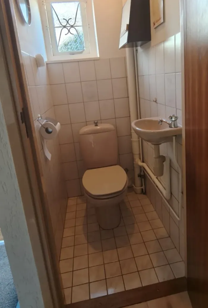 Vóór: smalle toiletruimte met beige staand toilet, fonteintje en wit tegelwerk onder een raampje met sierrooster